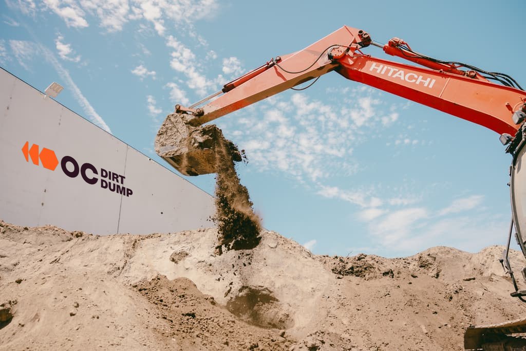 Excavator arm raised high, dirt falling from the bucket against a clear sky